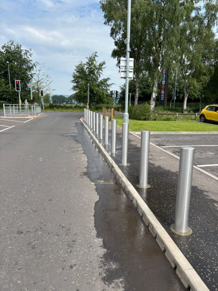 Completed stainless-steel bollards installed beside a retail car park access road in Bishopbriggs